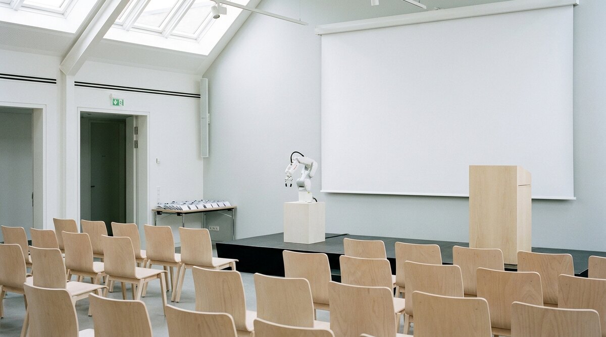 A bright hall set up before an audience arrives, rows of empty pale wooden chairs facing a low stage where a small white robot arm stands on a plinth beside a lectern, with a blank projection screen behind and a table of badges by the door